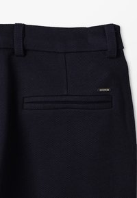 Pantalons en tissu texturé bleu marine avec une taille plate et une poche arrière unique ; comporte une étiquette de logo sur la taille.