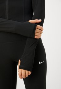 Svart idrottströja med långa ärmar, med tumhål och en tajt passform, kombinerad med svarta leggings som har en liten vit logotyp.