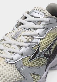 Κοντινή λήψη σε αθλητικό παπούτσι από λευκό και γκρι δίχτυ, με λευκά κορδόνια και μεταλλικό λογότυπο στο πλάι.