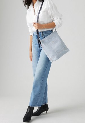 Světle modrá plátěná taška s proužky, přední kapsou, tmavým popruhem a červeným dekorativním šitím, nesená osobou v přiléhavých džínách a černých botách.