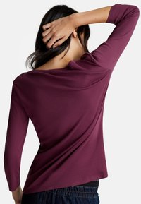 Haut à manches longues de couleur bourgogne foncé en tissu doux, avec un large col et un dos drapé, associé à un pantalon en jean foncé.