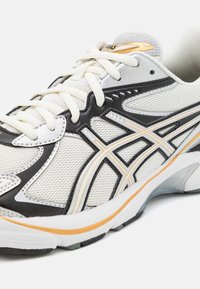 Vit träningsskor med mesh och syntetiska överlägg, utrustade med svarta och guldiga accenter, runda skosnören och en dämpad sula för stöd.