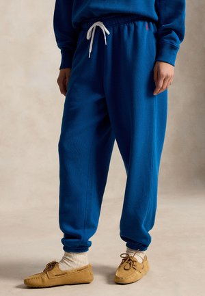 Άτομο που φοράει μπλε φόρμα με λευκό κορδόνι, μπεζ κάλτσες και καφέ μοκασίνια, στέκεται σε ουδέτερο φόντο.
