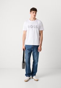 Witte katoenen T-shirt met de tekst "BOSS" in zilver; gecombineerd met blauwe denim jeans en witte sneakers. Man houdt een grijze schoudertas vast.