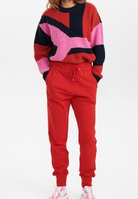 Person som bär röda joggerbyxor, en flerfärgad tröja med rosa, röd och svarta mönster, samt rosa-vita sneakers, står med händerna i fickorna.