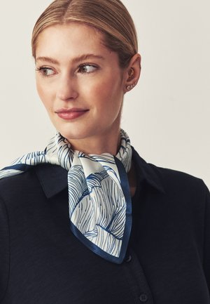 Femme blonde aux cheveux lissés en arrière porte une écharpe blanche et bleu marine à motifs et une chemise bleu marine à col, regardant légèrement à gauche avec une expression neutre.