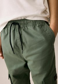 Pantalons cargo verts en mélange de coton. Caractéristiques : taille élastique, cordon de serrage noir, poches latérales et finition en tissu texturé.