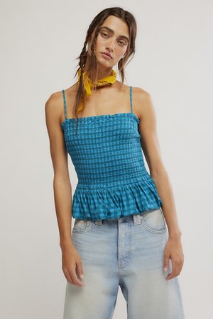 Kvinne med flettefrisyre, iført blå smokkert ermeløs topp med volangkant, lyse denimjeans og et gult halstørkle mot enkel bakgrunn.