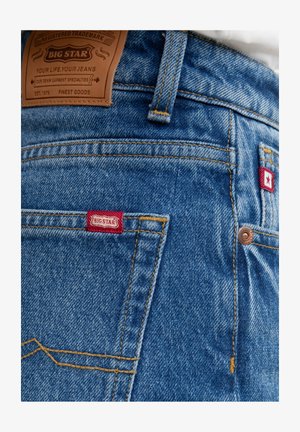 Blå denim jeans med en brun læderlap, røde mærkater, kobbernitter og gule syninger. Har en klassisk pasform og baglommer.
