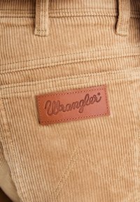 Tissu en velours côtelé tan avec une texture côtelée, présentant une étiquette en cuir marron embossée avec "Wrangler" et des détails de couture décoratifs.