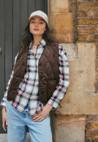 Eine Frau trägt eine beigen Mütze, eine braune Steppweste, ein kariertes Hemd und hellblaue Jeans und steht vor einer alten Ziegel- und Steinmauer.