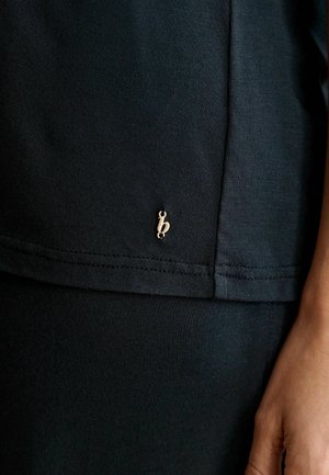 Tuva melna auduma apģērba gabala ar mazu zelta "b" logotipu pie apakšmalas un personas roka, kas atpūšas pie apģērba.