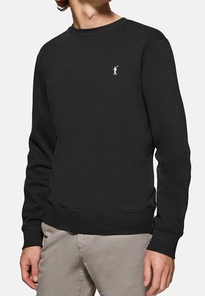 Man draagt een zwart crewneck sweatshirt met een klein wit golfspelerslogo op de linkerborst en beige broek.