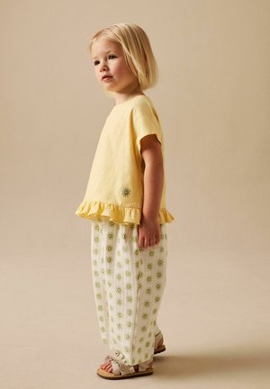 Copil tânăr cu păr scurt blond, îmbrăcat într-un top galben cu volane și pantaloni albi cu modele verzi de soare, sandale bej.