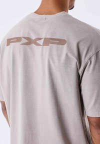 Camiseta de algodón gris con cuello redondo y mangas cortas, con un gran logo beige "PXP" impreso en la parte posterior. Textura suave.