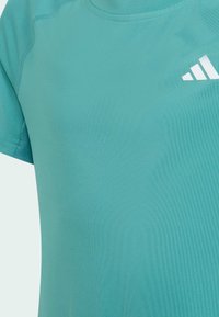 Koszulka sportowa z krótkim rękawem w kolorze teal, wykonana z miękkiej, przewiewnej tkaniny, z białym logo na górnej lewej stronie oraz delikatnym detalem teksturowym.