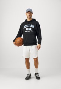 Felpa nera con la scritta "JORDAN" e "SPORT" in blu, pantaloncini bianchi, palla da basket in mano, cappellino grigio e scarpe sportive nere.