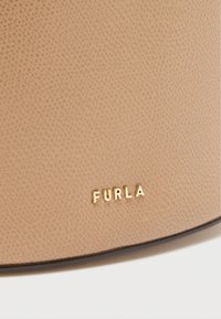 Béžová kožená taška s textúrovaným povrchom a zlatým ražbou logom "FURLA" na hladkej časti. Okraj je zdobený tmavým kontrastným lemovaním.