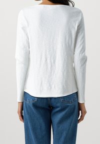 Maglietta bianca a maniche lunghe con scollo rotondo, realizzata in tessuto morbido. Abbinata a jeans in denim blu a vita alta, dotati di tasche posteriori.