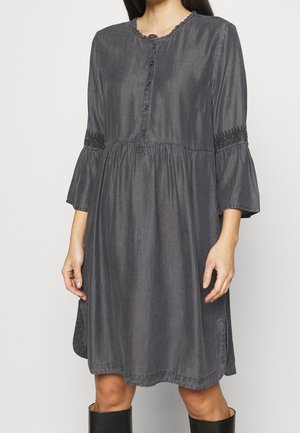 Femme portant une robe ample, gris foncé, boutonnée, avec des manches trois-quarts ornées de détails en dentelle noire au-dessus des coudes.