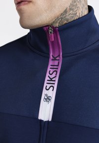 Giacca a zip blu navy con colletto alto e zip viola. Presenta una texture elegante e il marchio SIKSilK sul nastro della zip.