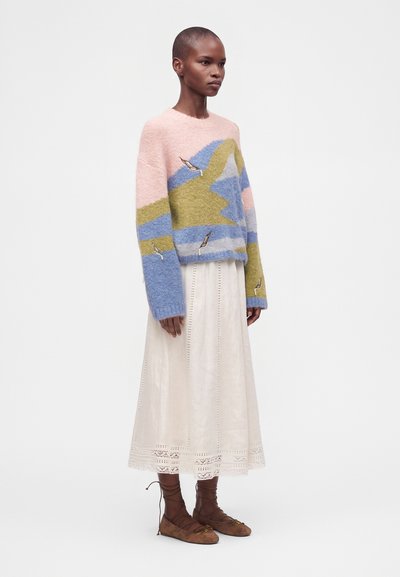 Femme aux cheveux courts portant un pull en laine doux à blocs de couleurs pastel, une longue jupe blanche avec bordure en dentelle, et des ballerines marron à lacets sur fond blanc.