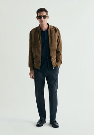 Hombre con gafas de sol oscuras, chaqueta bomber de ante marrón, camisa negra, pantalones negros y zapatos negros de pie contra un fondo claro.