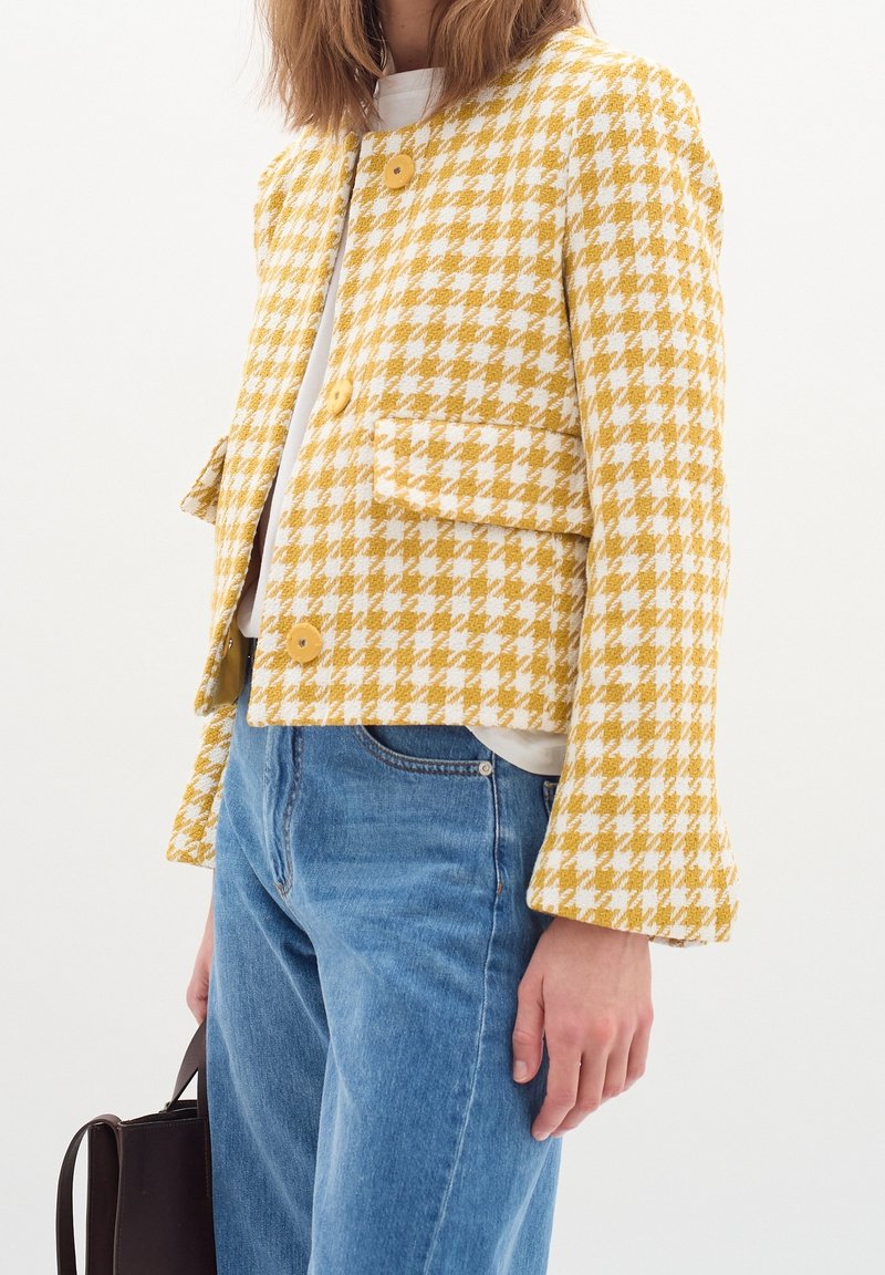 Personne portant une veste pied-de-poule jaune et blanche avec de gros boutons, jean bleu, chemise blanche, tenant un sac à main sombre sur le côté.