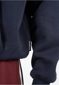 El sudadera de color navy presenta una textura acanalada, capucha y una etiqueta contrastante con la palabra "PROHIBIDO" mostrada en la costura lateral.