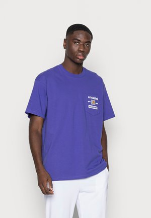 T-shirt de algodão roxa com mangas curtas, apresentando um bolso no peito e texto gráfico "scrambled" e "NETWORK" em cores contrastantes.