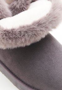 Zapatilla gris con un suave y esponjoso ribete de pelaje gris claro y un upper de textil liso. Presenta una forma redondeada y un interior acolchado.