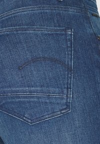 Mörkblå denimjeans med en bakficka som har en subtil böjd sömdekoration. Tyget har en lätt lyster och är mjukt i texturen.