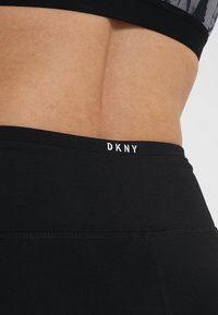 Černé sportovní legíny DKNY mají hladkou texturu a vysoký pás. Logo značky je viditelné v bílé barvě na pase.