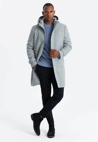 Abrigo gris claro con capucha, con cierres a presión. Usado sobre un suéter de canalé azul y pantalones oscuros a cuadros, combinado con botas negras de tobillo.