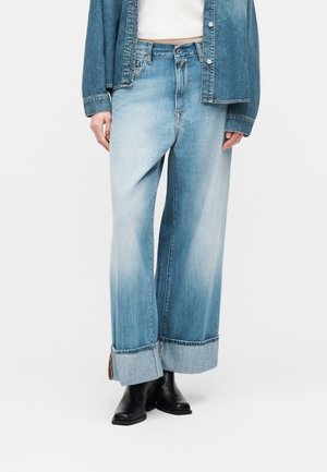 Άτομο που φοράει ανοιχτό μπλε τζιν παντελόνι με φαρδιά μπατζάκια και μεγάλες πιέτες, μαύρα μποτάκια μέχρι τον αστράγαλο, λευκό κοντό τοπ και ανοιχτό τζιν μπουφάν.