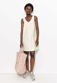 Robe sans manches jaune clair avec un décolleté en V, présentant un design texturé et des poches. Un sac fourre-tout à carreaux roses et des baskets argentées complètent le look.