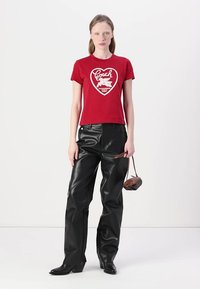 Camiseta de algodón roja con un gráfico en forma de corazón; combinada con pantalones de cuero sintético negros y un bolso marrón; la modelo está de pie contra un fondo blanco.