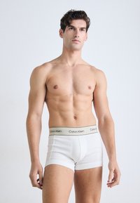 Bovenlichaamloze man met kort krullend haar, gekleed in witte boxer briefs met een beige Calvin Klein-broekband, staand tegen een effen lichte achtergrond.