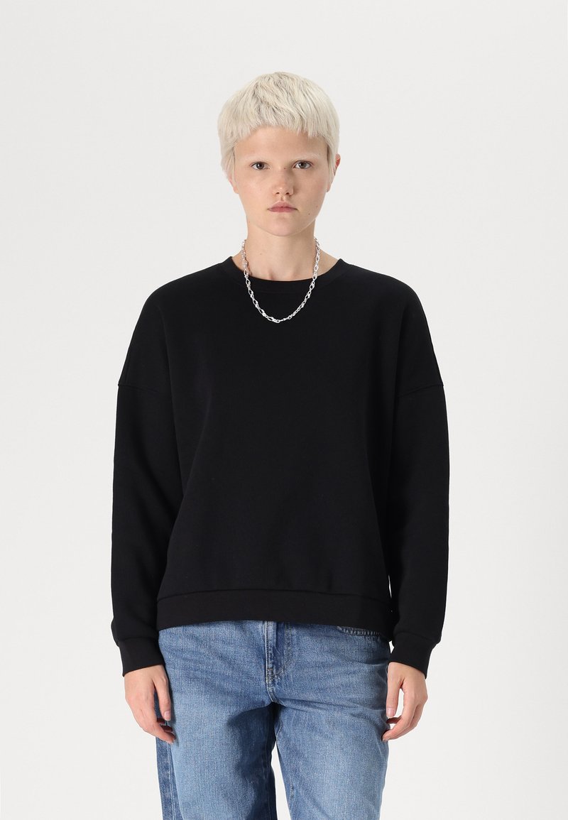 Sudadera negra de manga larga, cuello redondo y dobladillo de canalé. Combinada con jeans de color azul claro, tiene un peinado corto y un collar de cadena de plata.