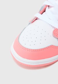 Valge tennisejalanõu roosade aksentidega, millel on sile nahast pealmine osa, aukudega varvaste osa hingavuse tagamiseks ja imitatiivse mustriga talla haarduvuse parandamiseks.