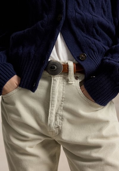 Personne portant un pantalon en velours côtelé beige avec les mains dans les poches, une ceinture en cuir marron avec une boucle argentée, une chemise blanche et un cardigan bleu marine.