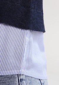 Navy textured knit top layered over a light blue and white striped shirt, partially tucked into light wash denim.