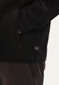 Black corduroy jacket with ribbed texture, side pockets, and a small logo patch. Worn over dark brown pants, highlighting the layered look.