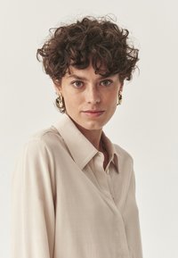 Camicia button-up beige con colletto, caratterizzata da una texture liscia e maniche lunghe. Orecchini dorati sono visibili, aggiungendo un accento metallico.