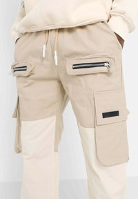 Beiga cargobyxor har dragkedjefickor på sidorna, en ficka med patch och en färgblockerad design med en jämn struktur och justerbar dragsko.