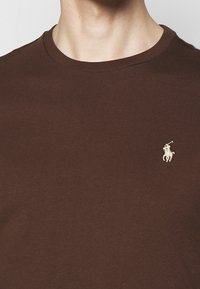 Hnědá bavlněná tričko s kulatým výstřihem. Má malé, vyšívané logo světle barvy na levé části hrudi. Hladká textura.