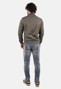 Giubbotto bomber verde con una texture liscia, abbinato a jeans chiari strappati con un logo sulla tasca posteriore e sneakers marroni.