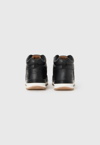 Pair of black mid-top sneakers with white midsoles and tan outsoles, seen from the back on a plain light background.
