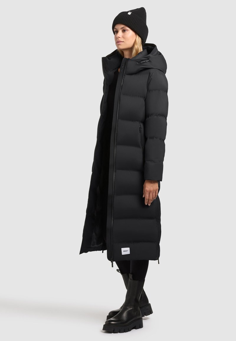 Cappotto puffo nero con un lungo design trapuntato, collo alto, zip frontale e tasche laterali. Indossato con un berretto nero e stivali neri spessi.
