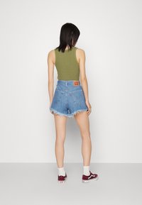 Top sin mangas verde acanalado, pantalones cortos de mezclilla azules con dobladillo deshilachado y zapatillas burdeos con cordones blancos, vistas desde la parte de atrás.